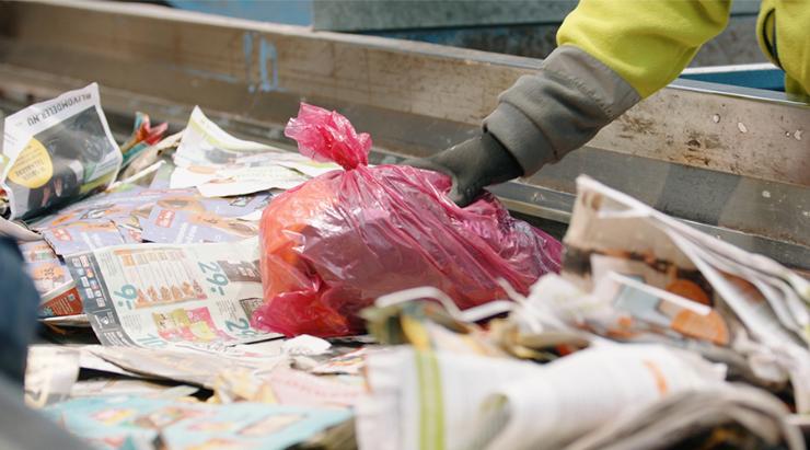 Person med gult arbejdstøj sorterer rød pose med tekstilaffald fra papiraffald på sorteringsanlæg